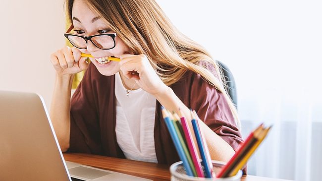 Mädchen mit Brille beißt auf einen Stift und sitzt vor einem Laptop